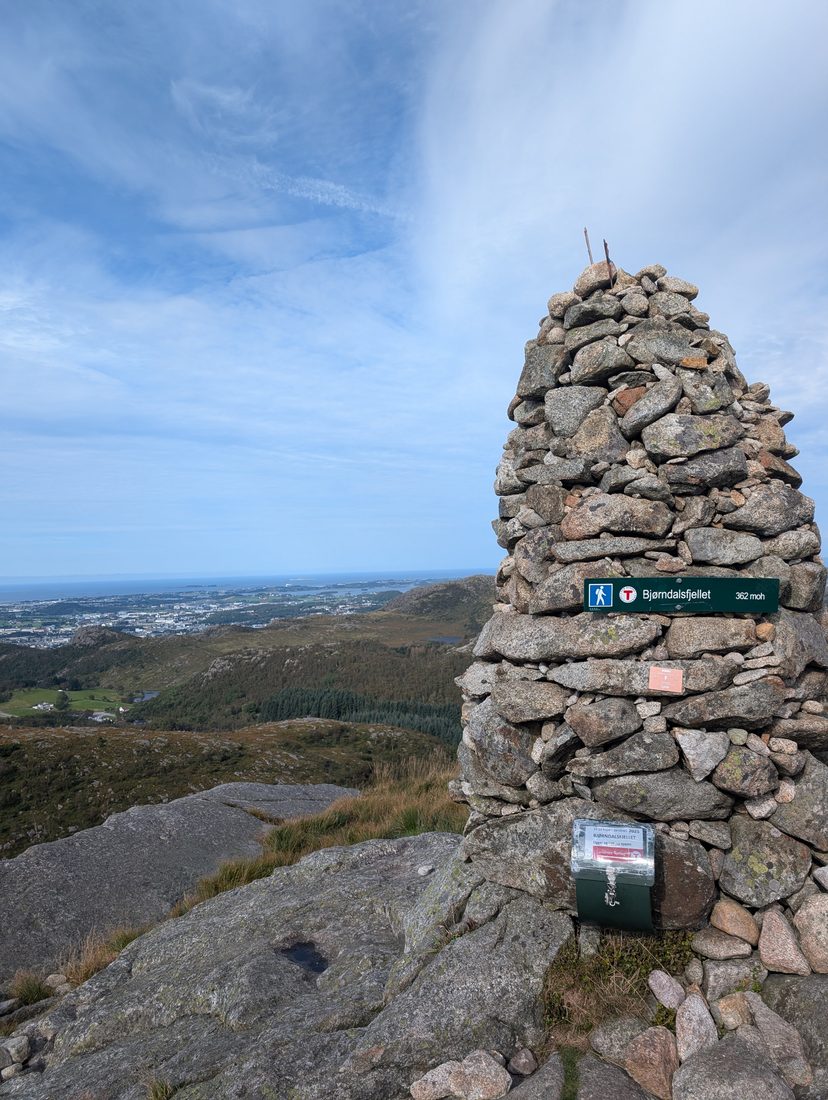 A stone trail cairn marking the summit of Bjørndalsfjellet, with a view over the coast below