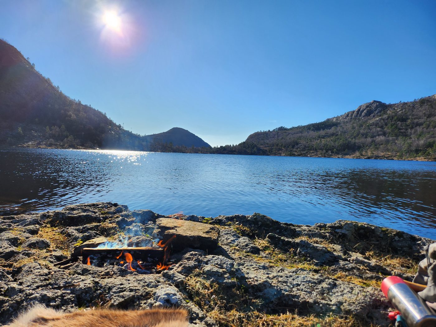 A campfire on a rocky lakeshore under a clear blue sky