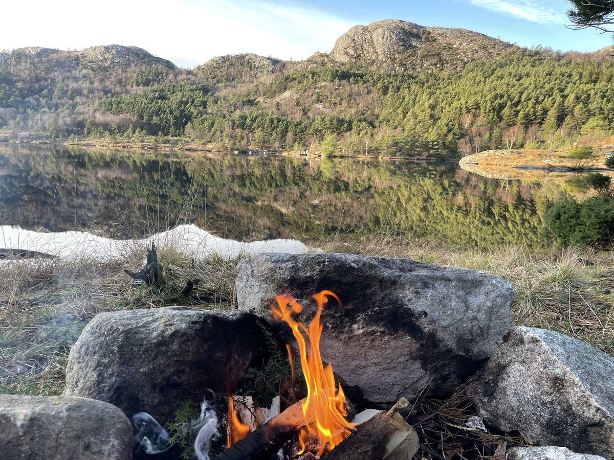 A still mountain lake reflecting the surrounding peaks, with a campfire in the foreground