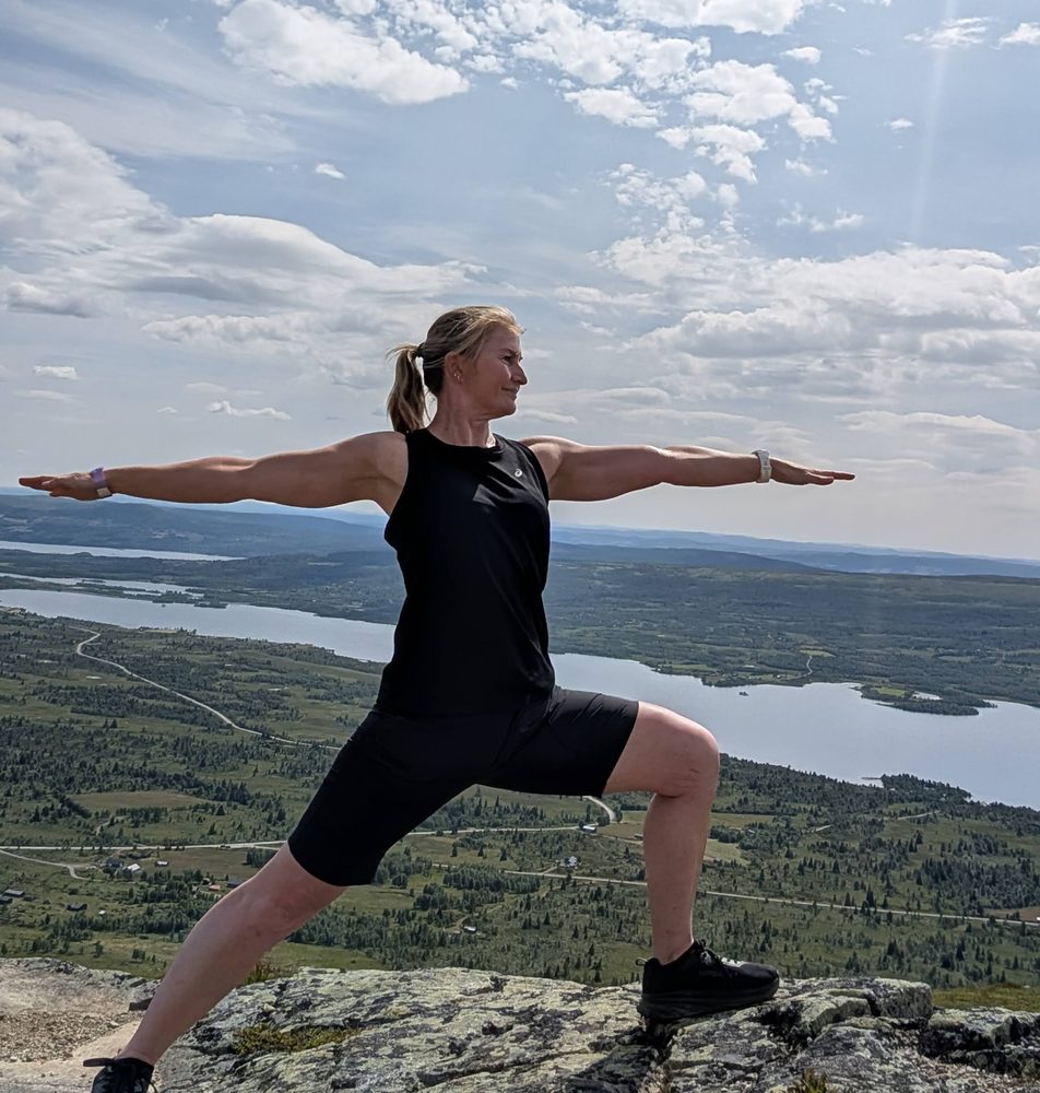Lena in a warrior pose on a mountain summit, overlooking a lake